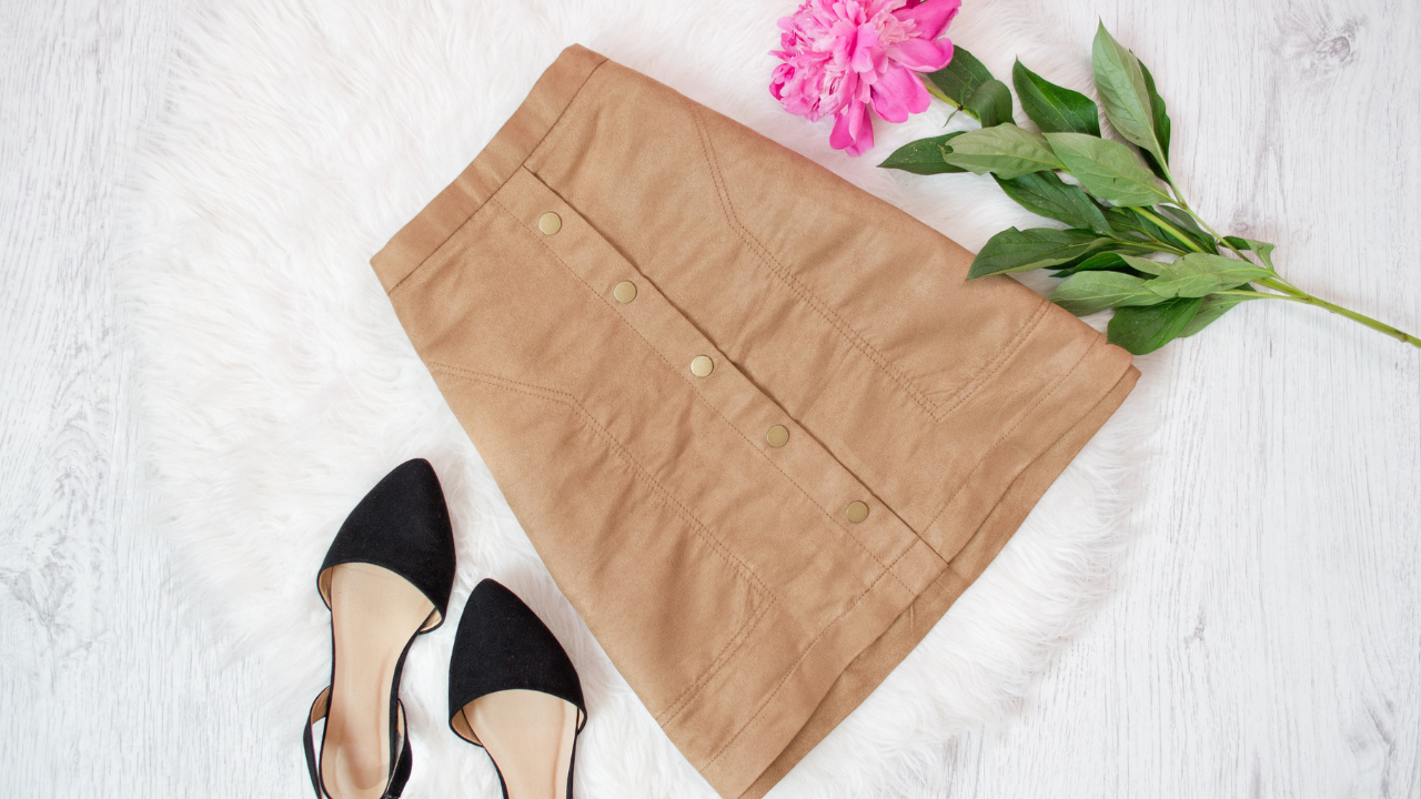 A tan button-front skirt, a classic among types of skirts, is laid on a white fur surface next to black pointed-toe flats and a pink flower with green leaves.